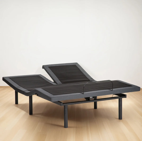 A wide-angle view of the Customatic Synergy Adjustable Base in a split-head configuration. The dark grey upholstered frame features two independent head sections, with one side inclined for sitting and the other remaining flat. The foot section remains a single unified piece with a black metal mattress retainer bar.