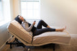 A happy woman is seen in a profile view, relaxing on the affordable Flexabed Value-Flex adjustable bed, which is raised at the head area. She is holding the remote control, demonstrating the bed's customizable features.