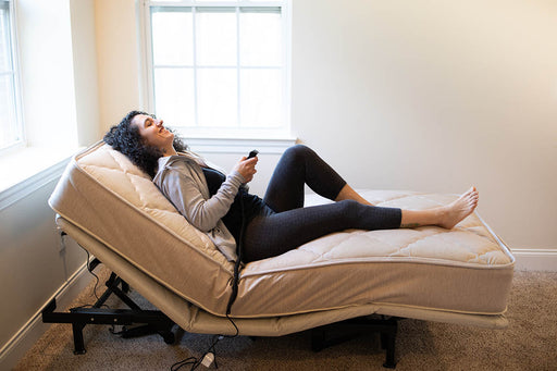 A happy woman is seen in a profile view, relaxing on the affordable Flexabed Value-Flex adjustable bed, which is raised at the head area. She is holding the remote control, demonstrating the bed's customizable features.