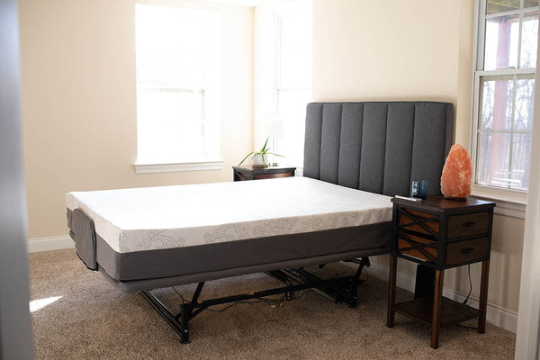 A modern adjustable bed in a bright, neutral-toned bedroom. The bed features a white mattress with a patterned top on a dark gray upholstered adjustable base, currently set in a flat position. The head of the bed is backed by a tall, vertical channel-tufted dark gray headboard. To the right is a rustic-industrial style nightstand with a dark wood top, two drawers, and an open shelf, featuring a large Himalayan salt lamp, a glass, and a remote control on top.