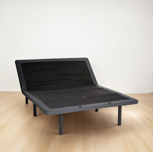 A front-facing three-quarter view of the Customatic Synergy Adjustable Base. The head section is partially raised, showcasing the smooth transition between the dark grey fabric upholstery and the black non-slip grip material on the surface.