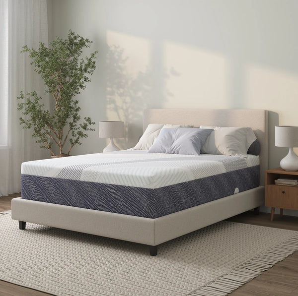 A modern bedroom scene featuring the DreamTemp mattress on a beige upholstered bed frame. The room is brightly lit with a large potted plant and minimalist nightstands, highlighting the mattress’s stylish blue and white profile.