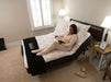 A woman in light pink pajamas sits propped up in the Flexabed Premier adjustable bed, reading a book. The head of the bed are raised to a comfortable incline, showcasing the bed's customizable positions