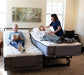 A man and a woman relax in a split Flexabed Premier adjustable bed setup. The man is reading on his side, which is inclined, while the woman is lying down on her side of the bed. A caretaker is on the right side of the bed, seemingly adjusting the woman's pillow