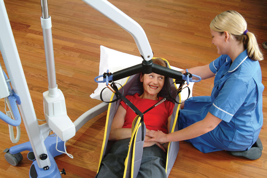 A nurse in blue scrubs smiles while assisting a woman lying in a transfer sling attached to the Joerns Hoyer Advance Electric Patient Lift—Compact Folding Floor Lift, on a wooden floor. The woman looks comfortable and gazes up at the nurse during the transfer.