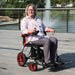 A woman with glasses and a floral shirt sits smiling in a Journey Air Elite power chair on an outdoor boardwalk. The power chair features vibrant red accents on the armrests and wheel rims, with a scenic park and fountain in the background.