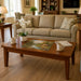 A wooden rectangular coffee table in a warm brown finish, styled in a cozy living room with a beige sofa, potted white orchids, mugs, and a colorful landscape jigsaw puzzle on top.