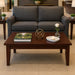 A dark brown wooden coffee table in a modern living room setting, styled with a book, a gold geometric ornament, a white candle, and a small glass vase with a white rose, positioned in front of a gray fabric sofa.