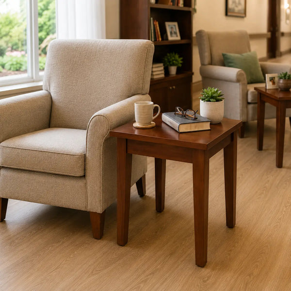 A square wooden end table with a warm medium-brown finish stands beside a beige upholstered armchair in a bright living room. On the tabletop are a white mug, a book, eyeglasses, and a small potted succulent.
