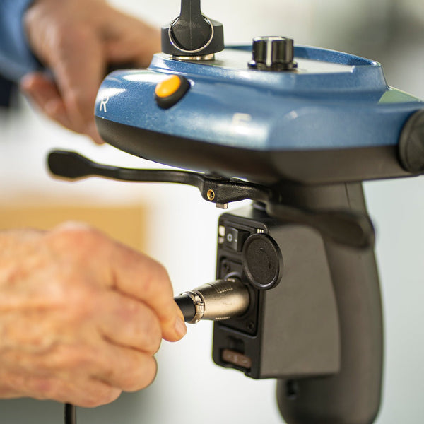 A close-up shot showing a person's hand plugging the charger into the port located on the tiller, just below the control panel. The image also shows the speed dial, yellow horn button, and the directional throttle levers.