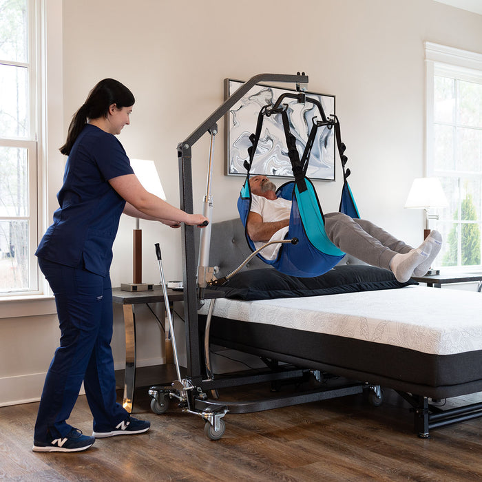 Caregiver using a Hoyer lift to assist a man in a sling above a Flexabed Hi-Low adjustable bed with optional mattress, demonstrating safe patient transfer