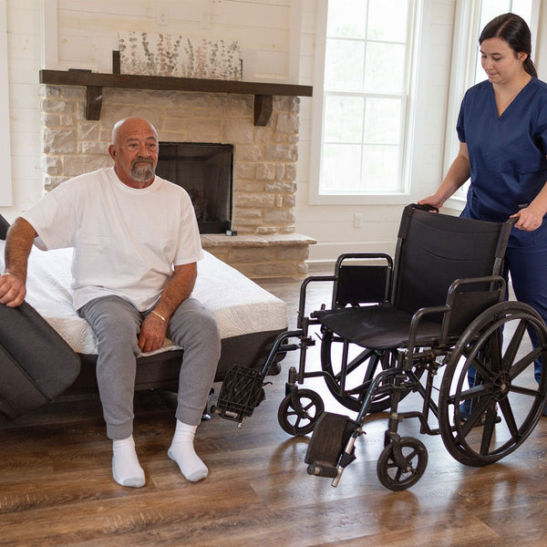 Older man sitting at the edge of a Flexabed Hi-Low adjustable bed with optional mattress, preparing to transfer into a wheelchair with caregiver assistance