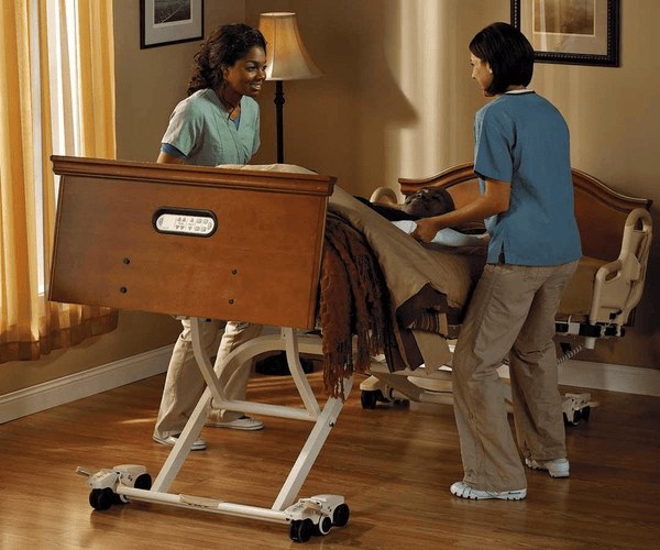 Two caregivers assisting a patient on the UltraCare XT bed system. The bed is tilted into the Gravity Assist position, showing the bed frame lifting and tilting for easier patient repositioning.