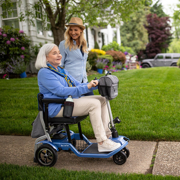 An elderly woman riding a blue Journey So Lite folding mobility scooter outdoors on paved path, accompanied by a younger woman.