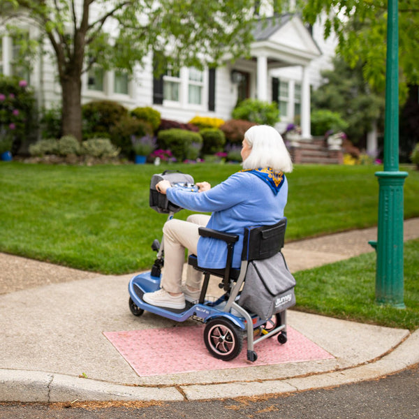 An elderly woman riding a blue Journey So Lite folding mobility scooter outdoors on the sidewalk.