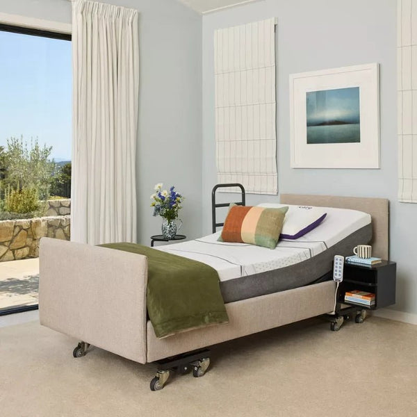 An iCare IC333 Luxury Electric Hospital Bed with low height access, dressed with pillows and a green throw, sits in a bright bedroom with large windows, beige carpet, and artwork. A nightstand holds flowers, a phone, and a remote control.