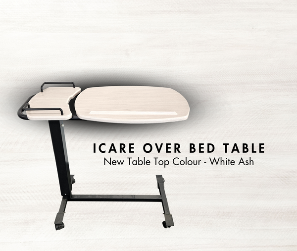 Promotional image for an Icare over bed table with a light-colored "White Ash" finish. The table is placed against a light wood-grain background. Text reads: "ICARE OVER BED TABLE. New Table Top Colour - White Ash."