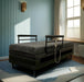 The UPbed Independence Adjustable Bed for Seniors by Journey Health, featuring black rails and a remote control, is placed in a sunlit room with blue paneled walls, a large curtained window, and a blue rug on the floor.