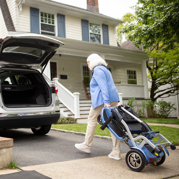 An elderly woman pulling a folded blue Journey So Lite mobility scooter like luggage outdoors on paved path, heading towards an SUV with an open trunk.
