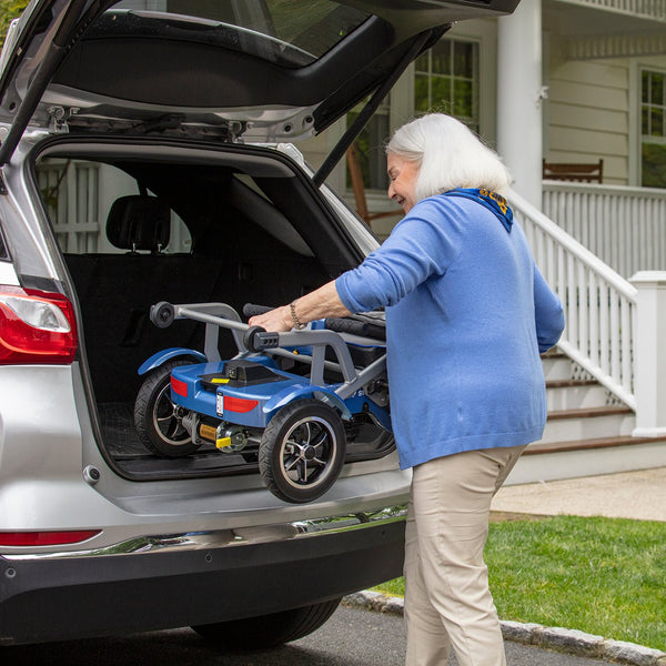 An elderly woman easily loading a folded blue Journey So Lite mobility scooter on a trunk of an SUV.