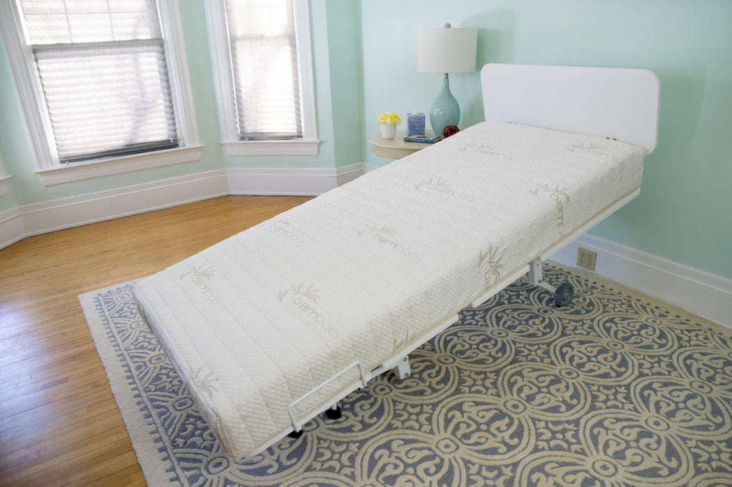 The Transfermaster Valiant Bariatric Hospital Bed with Trendelenburg and a white mattress is displayed in a modern room with light blue walls, wood floors, windows with blinds, a decorative rug, and a nearby nightstand with lamp. The head section is elevated.