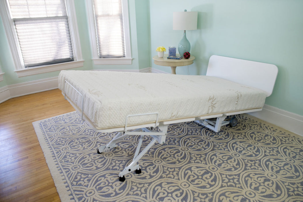 The Bariatric Hospital Bed with Trendelenburg - Transfermaster Valiant features an adjustable raised backrest and offers comfortable medical-grade positioning in a light blue room with hardwood floors, patterned rug, windows, and a small round table.