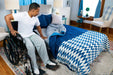 A man in a wheelchair sits beside the TransferMaster Bariatric Bed | Night Rider (Twin, Full, Queen) with blue bedding in a cozy, sunlit bedroom, preparing to transfer onto the bed.
