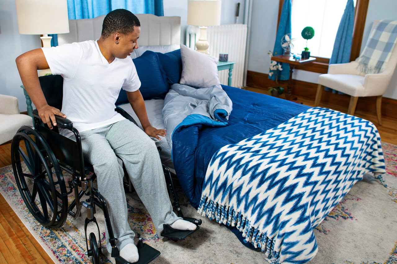 A man in a wheelchair sits beside the TransferMaster Bariatric Bed | Night Rider (Twin, Full, Queen) with blue bedding in a cozy, sunlit bedroom, preparing to transfer onto the bed.