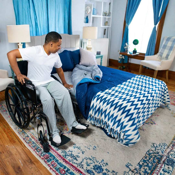 A man in a wheelchair sits beside the TransferMaster Bariatric Bed | Night Rider (Twin, Full, Queen) with blue bedding in a cozy, sunlit bedroom, preparing to transfer onto the bed.