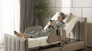 An older woman in pajamas relaxes on the Starsleep Orin Rotating Bed with its elevated backrest, using her smartphone. The bed includes a remote control, a nearby green plant, and soft daylight streaming through the window.