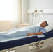 A middle-aged man rests comfortably on his back in a hospital bed covered by a light blue blanket, lying on the dark blue Proactive Protekt HC 50 foam mattress with cover. Bright sunlight streams in from a window on the left