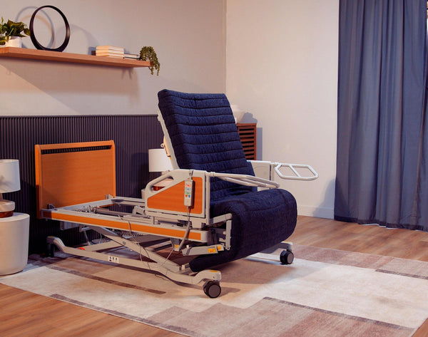 The ReNuCare Standing Bed, a 4-position adjustable hospital bed with a padded dark blue mattress and wooden accents, is set in a cozy, well-lit room featuring wooden flooring, shelving, and blue curtains.