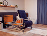 The ReNuCare Standing Bed, a 4-position adjustable hospital bed with a padded dark blue mattress and wooden accents, is set in a cozy, well-lit room featuring wooden flooring, shelving, and blue curtains.