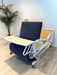 The ReNuCare Standing Bed is a 4-position adjustable hospital bed with a navy blue padded mattress, raised backrest, and attached light wood tray table, shown in a bright room with light wood flooring and a potted plant.