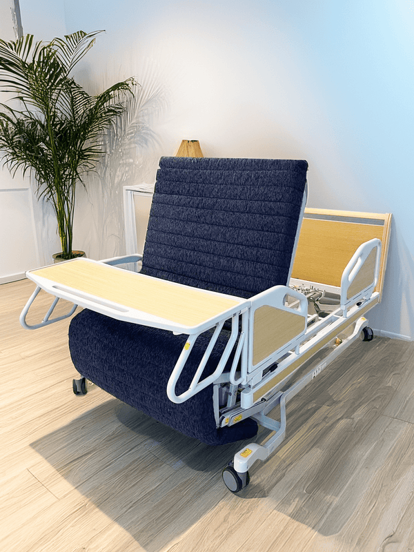 The ReNuCare Standing Bed is a 4-position adjustable hospital bed with a navy blue padded mattress, raised backrest, and attached light wood tray table, shown in a bright room with light wood flooring and a potted plant.