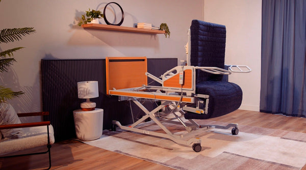 The ReNuCare Standing Bed—an adjustable 4-position hospital bed with wooden accents and wheels—stands in a cozy room featuring wooden floors, a blue velvet chair, plants, and a wall shelf with books and decor.