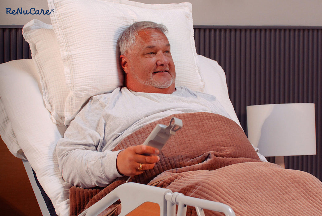 An older man with gray hair and beard lies in a hospital bed, smiling and holding a remote. Covered with a brown blanket and supported by white pillows, he uses the ReNuCare Standing Bed—4-position Adjustable. Logo appears in the top left corner.
