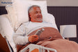 An older man with gray hair and beard lies in a hospital bed, smiling and holding a remote. Covered with a brown blanket and supported by white pillows, he uses the ReNuCare Standing Bed—4-position Adjustable. Logo appears in the top left corner.