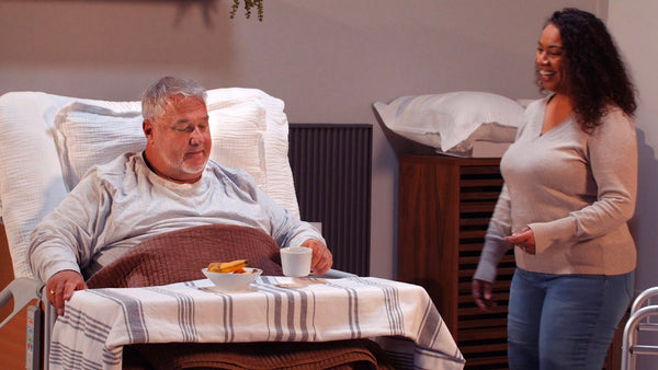 An older man sits up in a ReNuCare Standing Bed - 4-position Adjustable hospital bed with a tray of food and drink, while a smiling woman stands nearby in the warmly lit, comfortable room.