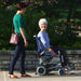 A lifestyle photo of two women outdoors on a paved path. An older woman with white hair is smiling while seated in a blue Journey Zinger, while a younger woman walks beside her.