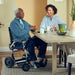 An elderly man sits in a Zoomer Power Chair at a wooden dining table, engaged in conversation with a woman. This demonstrates the chair's ability to fit comfortably under standard tables, functioning much like a regular chair in a home environment.