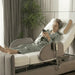 A senior woman relaxing in the Orin Star Sleep rotating medical bed. She is sitting up comfortably using her smartphone while the headrest is elevated. The image showcases the bed's modern grey upholstery, plush side rails, and dual wired remote controls in a home bedroom setting.