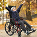 A young boy in a blue jacket and beanie sitting in a red Ziggo Pro wheelchair outdoors, laughing with his arms raised in a "V" sign to demonstrate the chair's outdoor mobility and user comfort.