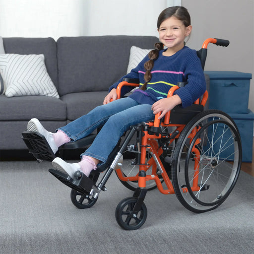 The young girl is shown in a living room setting, seated comfortably in the orange Ziggo wheelchair. In this view, the wheelchair is configured with elevating leg rests that are raised, supporting her legs while she relaxes near a sofa.