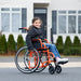 A young girl wearing a black winter jacket and jeans is seated in an orange Ziggo wheelchair on an outdoor paved surface. She is looking at the camera with a smile, demonstrating the chair’s use for community mobility in front of a residential home.