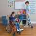 A young girl with long brown hair sits smiling in an orange Ziggo wheelchair in a brightly colored classroom. She is holding a clipboard and pen, engaged in schoolwork, while another student stands nearby using a blue pediatric posterior walker.