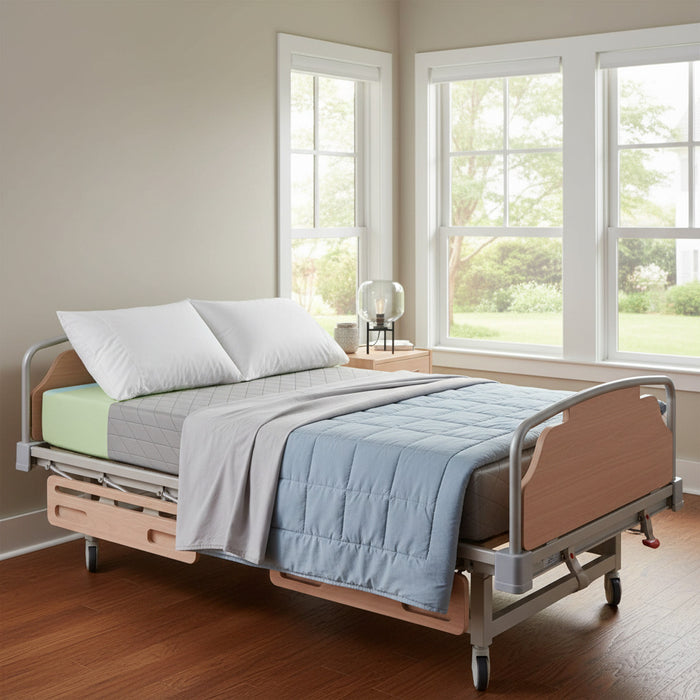 54" Wide Emerald Comfort Select Bariatric Mattress in a modern, residential-style bariatric hospital bed. A blue quilted blanket is draped on the mattress. The bed is shown in a brightly lit room with large windows.