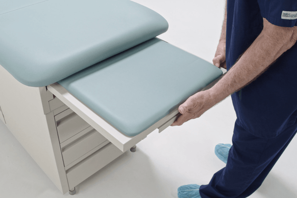 A close-up of a healthcare professional extending the padded leg rest extension from the foot of the Hausted 4200 Exam Table. The extension slides out smoothly to provide additional surface area for supine examinations.