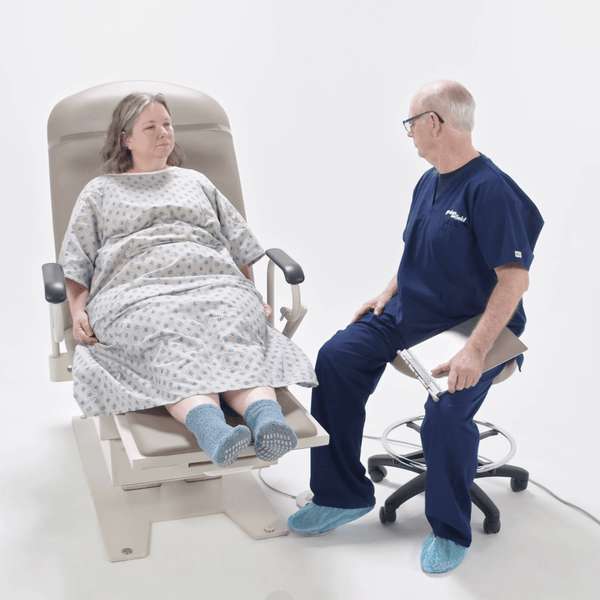 A female patient in a medical gown sits comfortably on the Graham-Field Hausted 4600 Series Powered Exam Table while a healthcare professional sits nearby on a rolling stool. The table is shown in a neutral tan upholstery, featuring a low-access height design, integrated armrests for patient security, and a powered adjustable backrest and leg section that allows the patient to remain seated during the consultation.