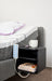 The bedside shelf unit is shown attached to a grey upholstered Icare bed. It holds a white coffee mug and eyeglasses on top, while the lower shelves store a pen and books (including "The Self-Care Journal").